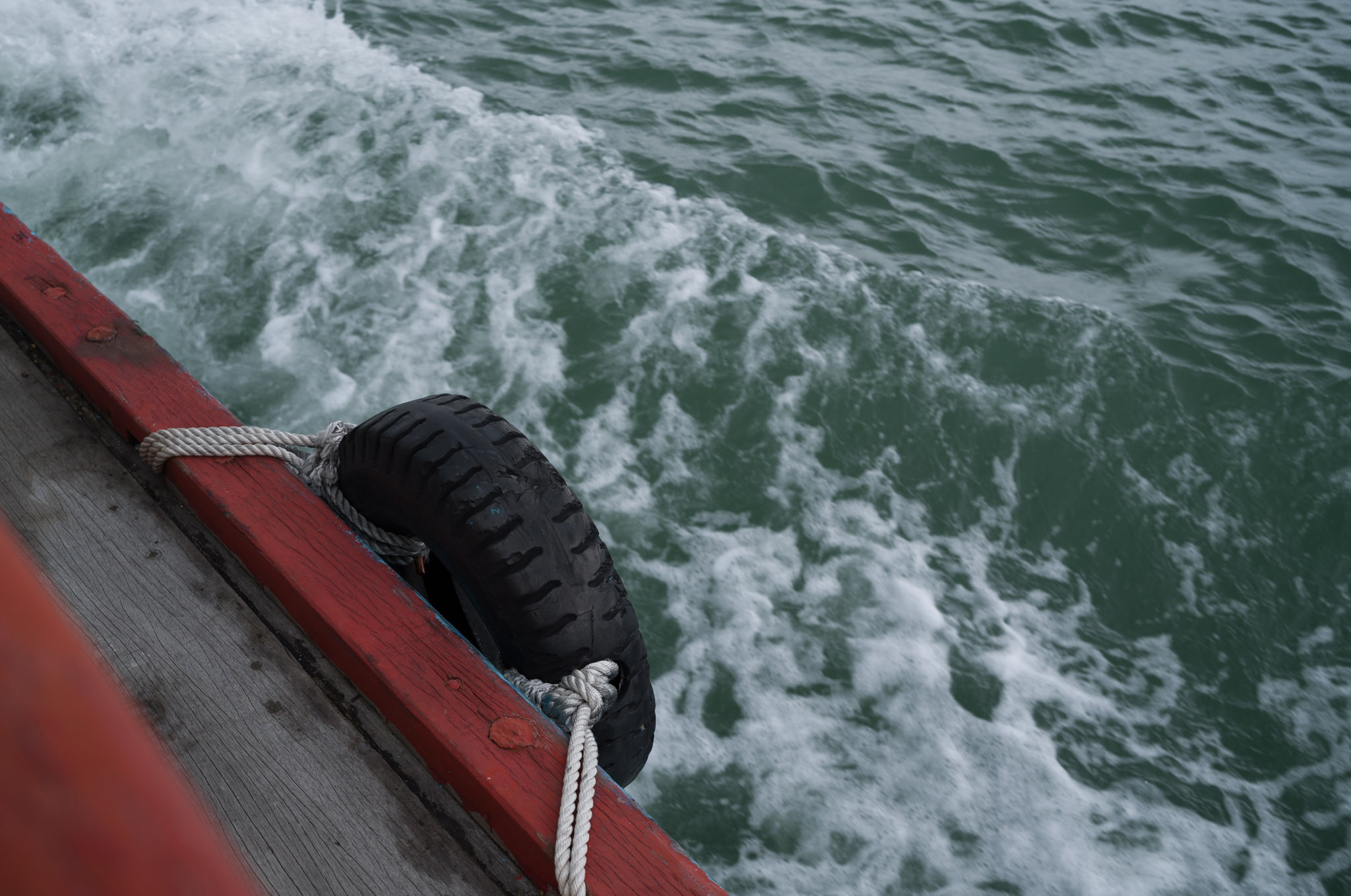 A rubber tyre fender roped to the red wooden hull of a bumboat, with churning green wake behind.