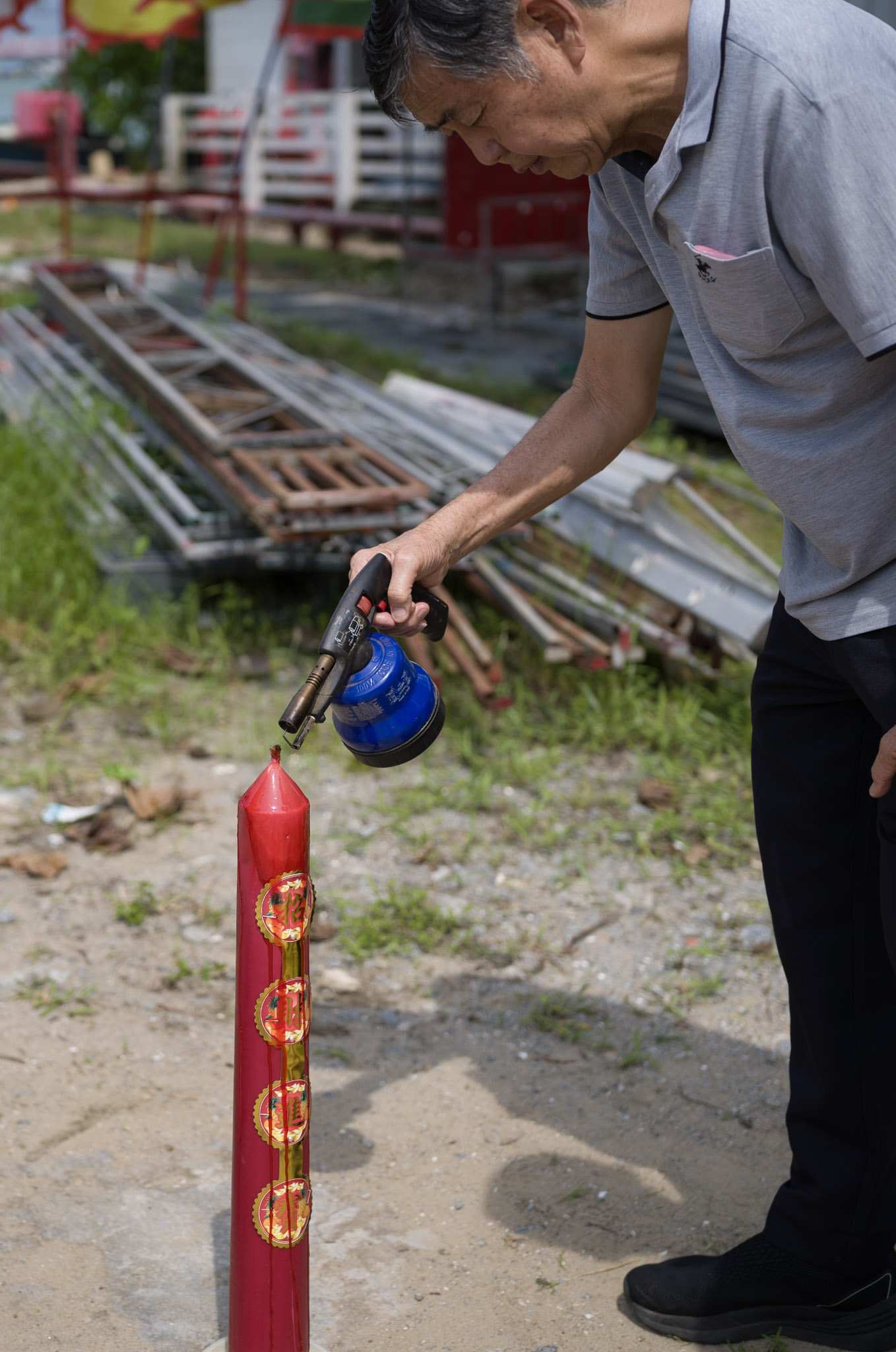 A man bends over a tall red ceremonial candle decorated with gold medallions, lighting it with a blowtorch.