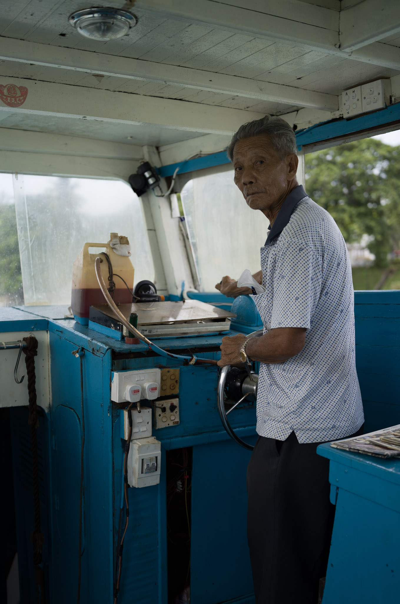 An elderly man stands at the helm of a blue-painted bumboat cabin, looking back at the camera.