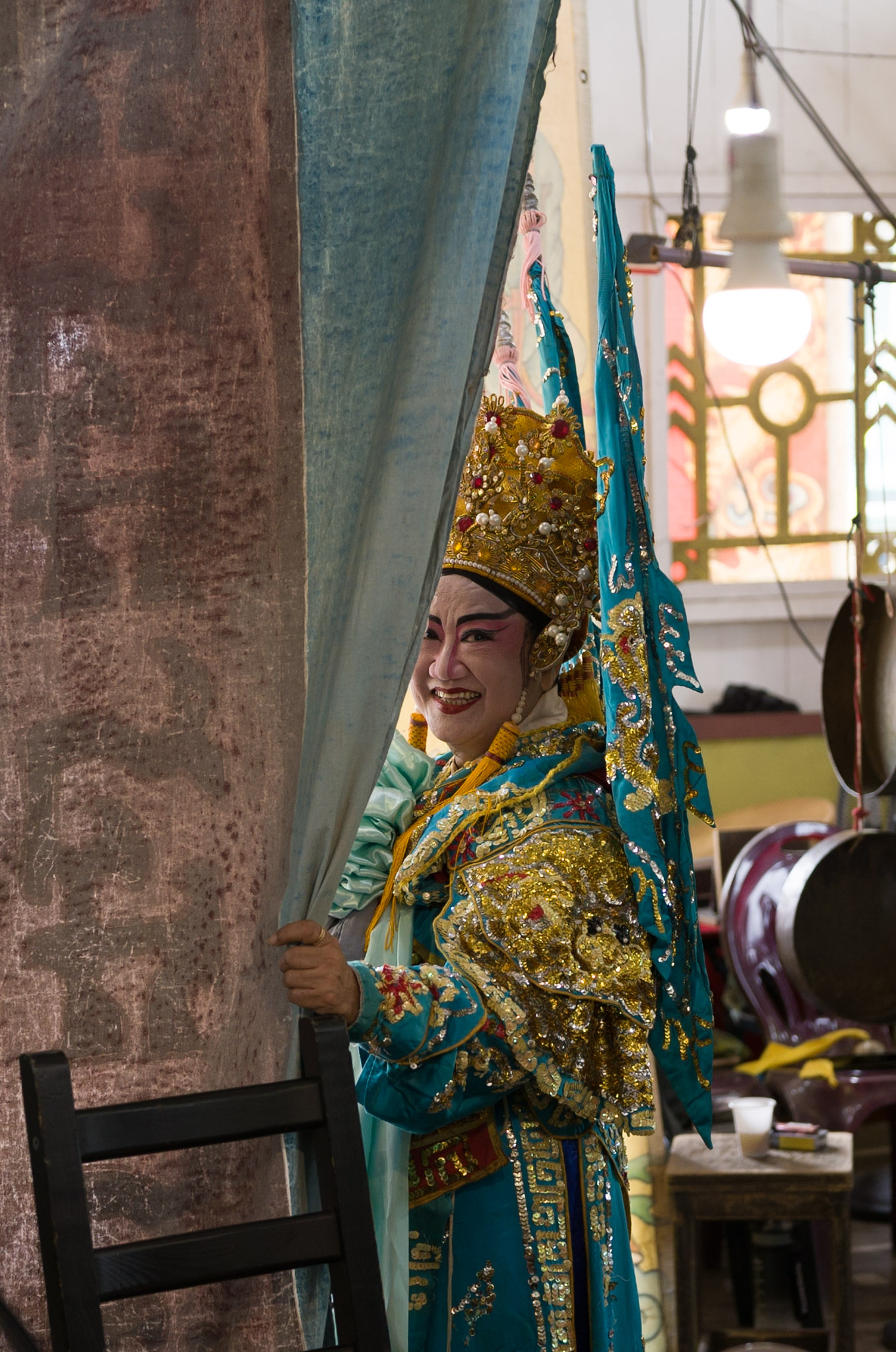 A performer in a teal and gold opera costume with headdress smiles while pulling back a curtain to peek at the stage, drum and gong visible behind.