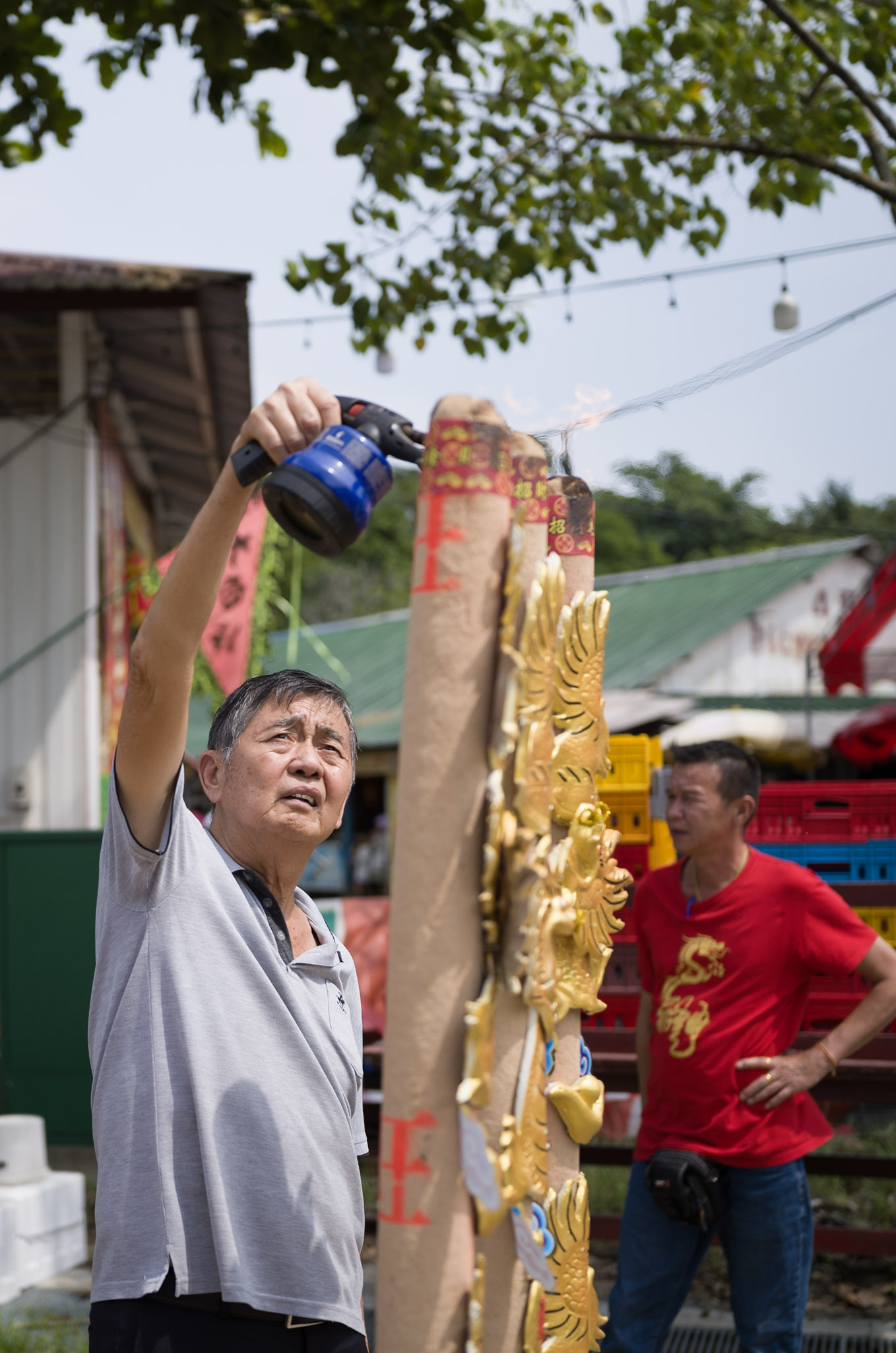 A man reaches up to light a tall paper-wrapped incense column beside a gold dragon column with a blowtorch, a second man in a red shirt watching.