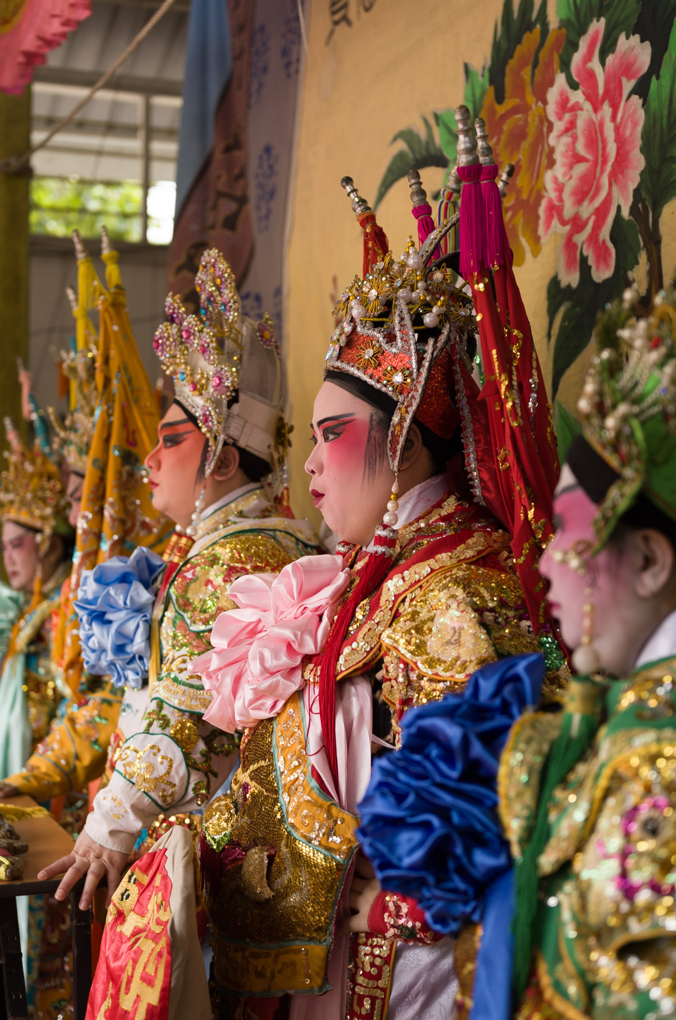 Five opera performers in full imperial costumes and jewelled headdresses stand in profile in the wings, waiting for their entrance.