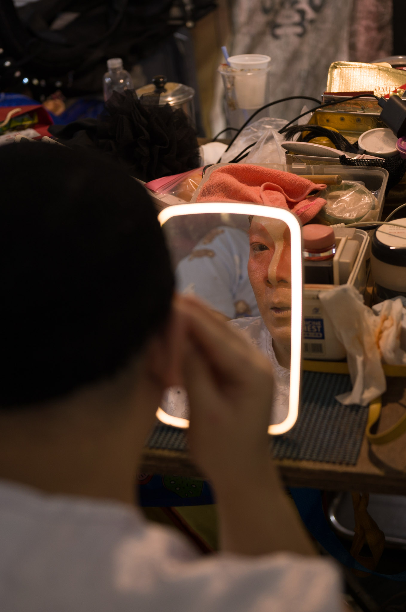 A performer seen from behind applies white face paint, their reflection visible in a glowing lit vanity mirror on a cluttered dressing table.