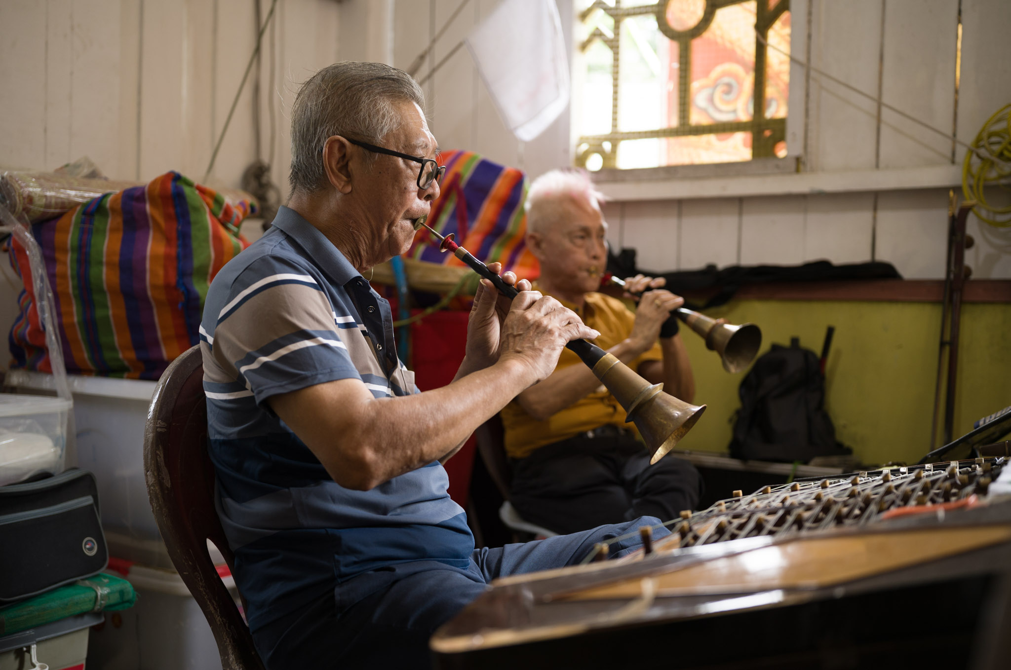 Two elderly men play suona wind instruments side by side in a cluttered backstage room, a mixing board in the foreground.