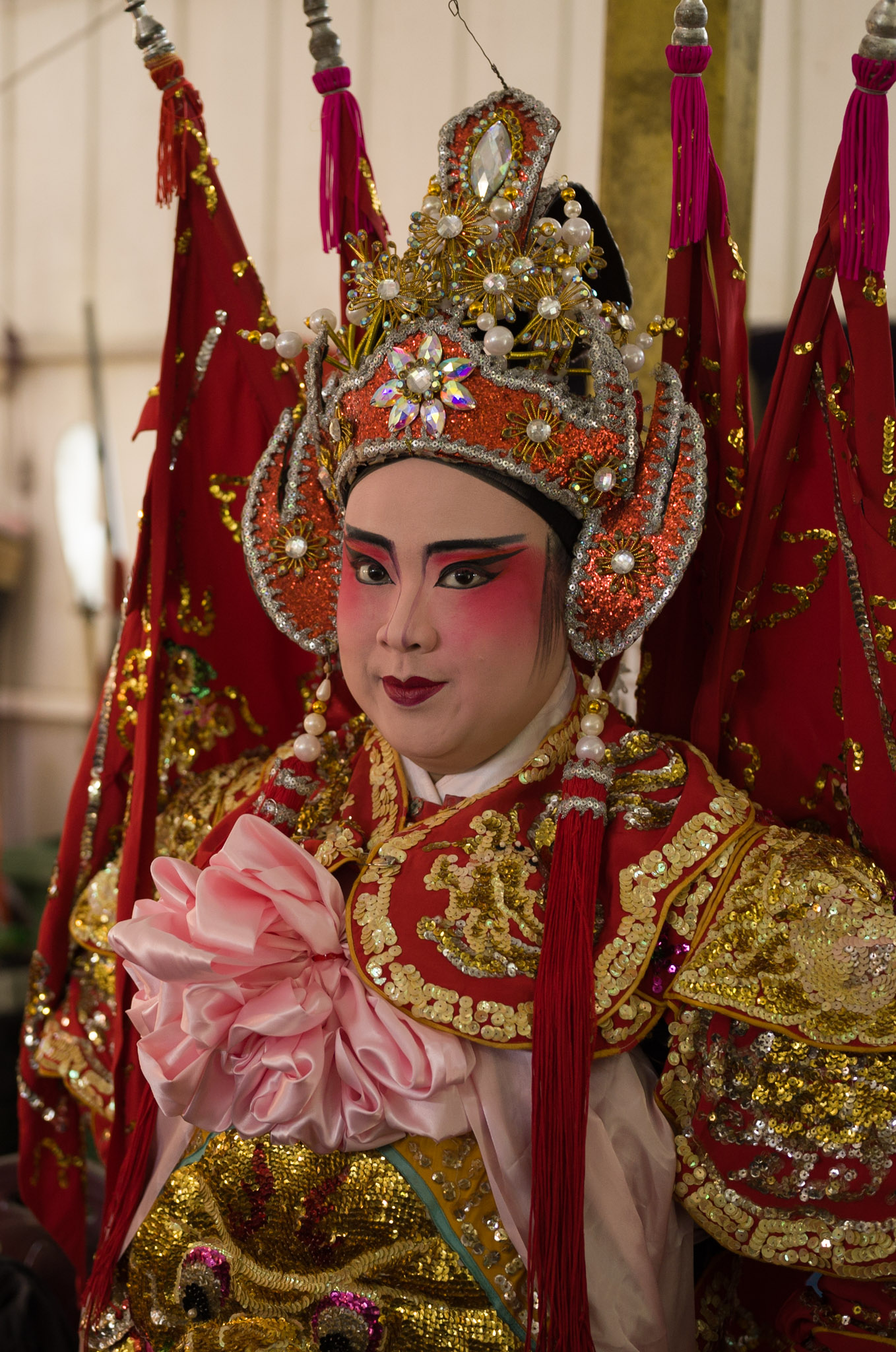 A performer in a red and gold imperial opera costume with jewelled headdress holds a pink flower prop, flags fanning behind.