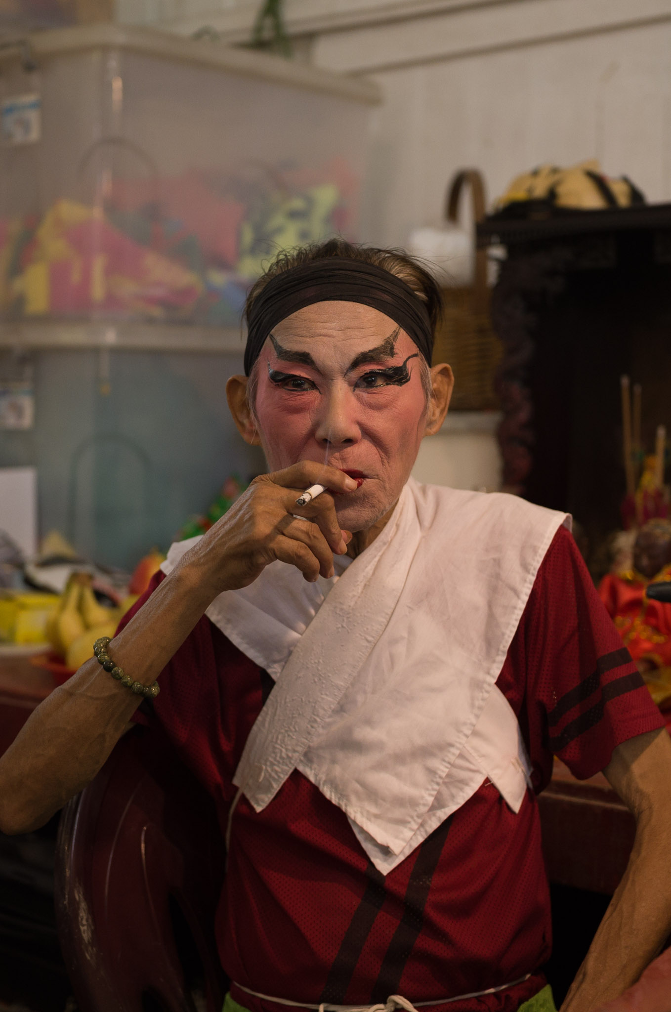 An opera performer in white and dark demon face paint smokes a cigarette backstage, a white towel draped around his neck.
