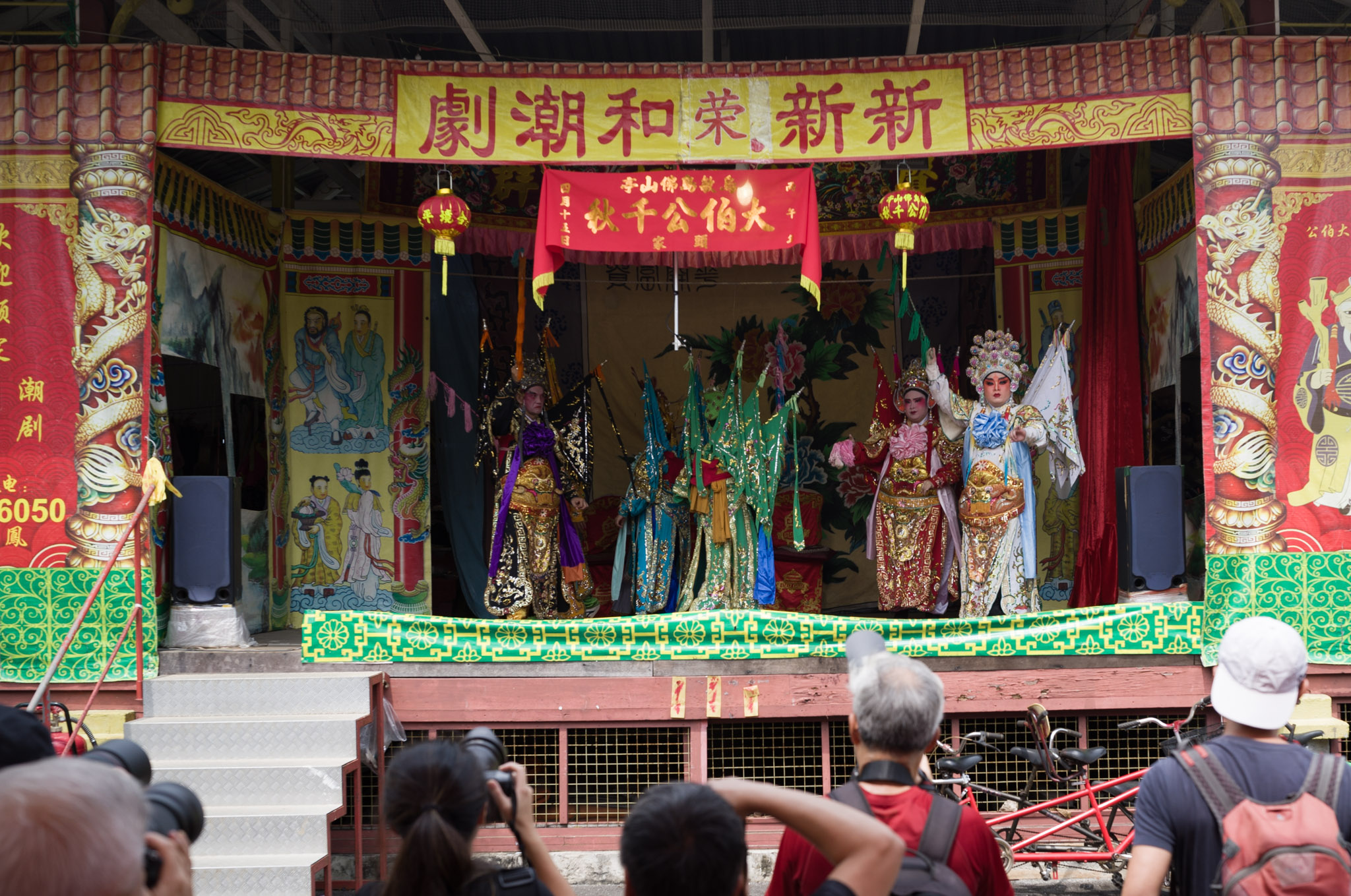 The ornate Xin Xin Rong He Teochew opera stage with red and gold proscenium, performers mid-scene under a large yellow banner, watched by a crowd below.