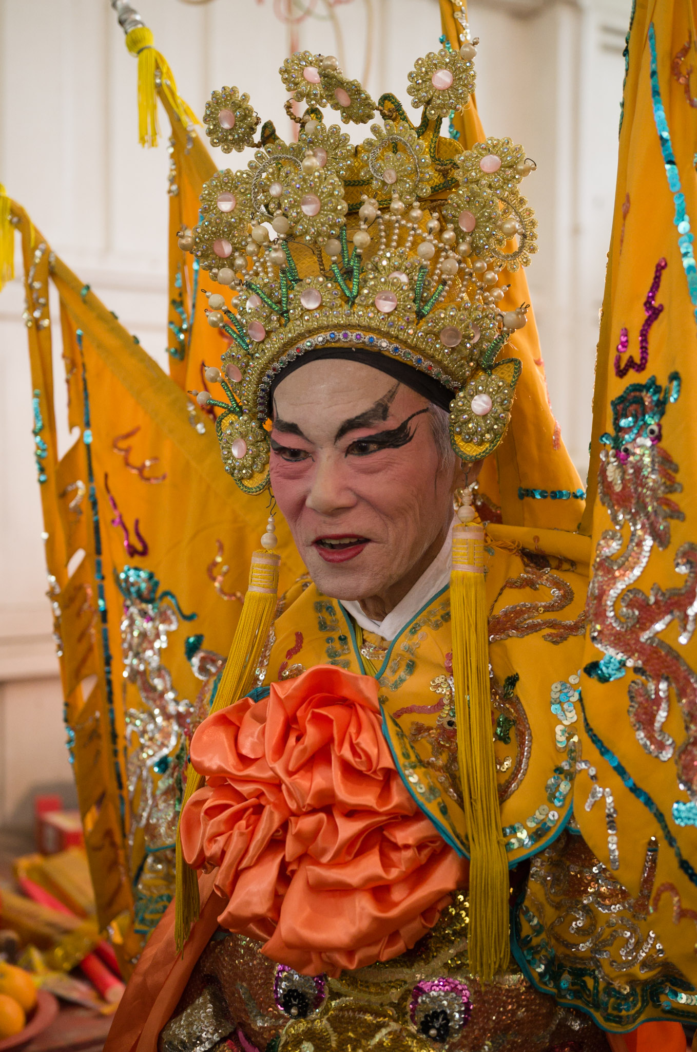 A performer in a yellow and gold imperial opera costume with tall jewelled headdress holds an orange flower, yellow flags behind.